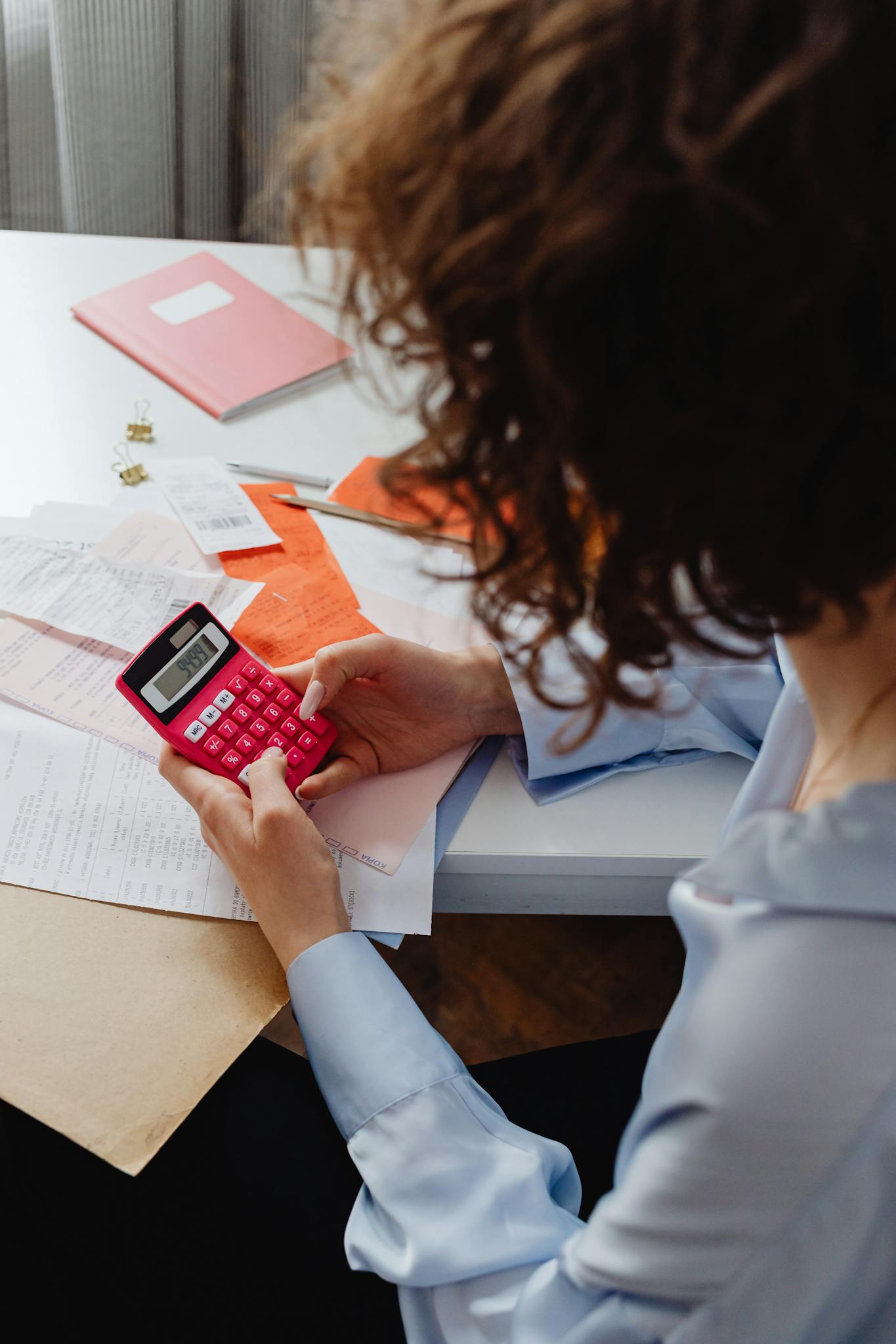 Adult woman working with receipts and a bright pink calculator at a desk, focusing on finances.